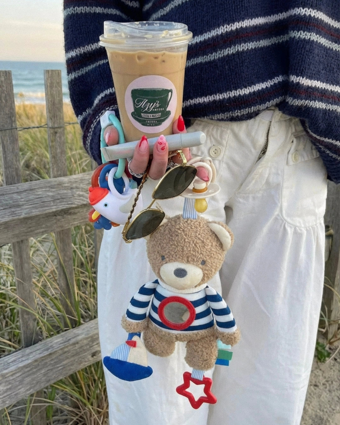 Person holding a cup with a teddy bear keychain, standing by a wooden fence with grass and sky in the background.