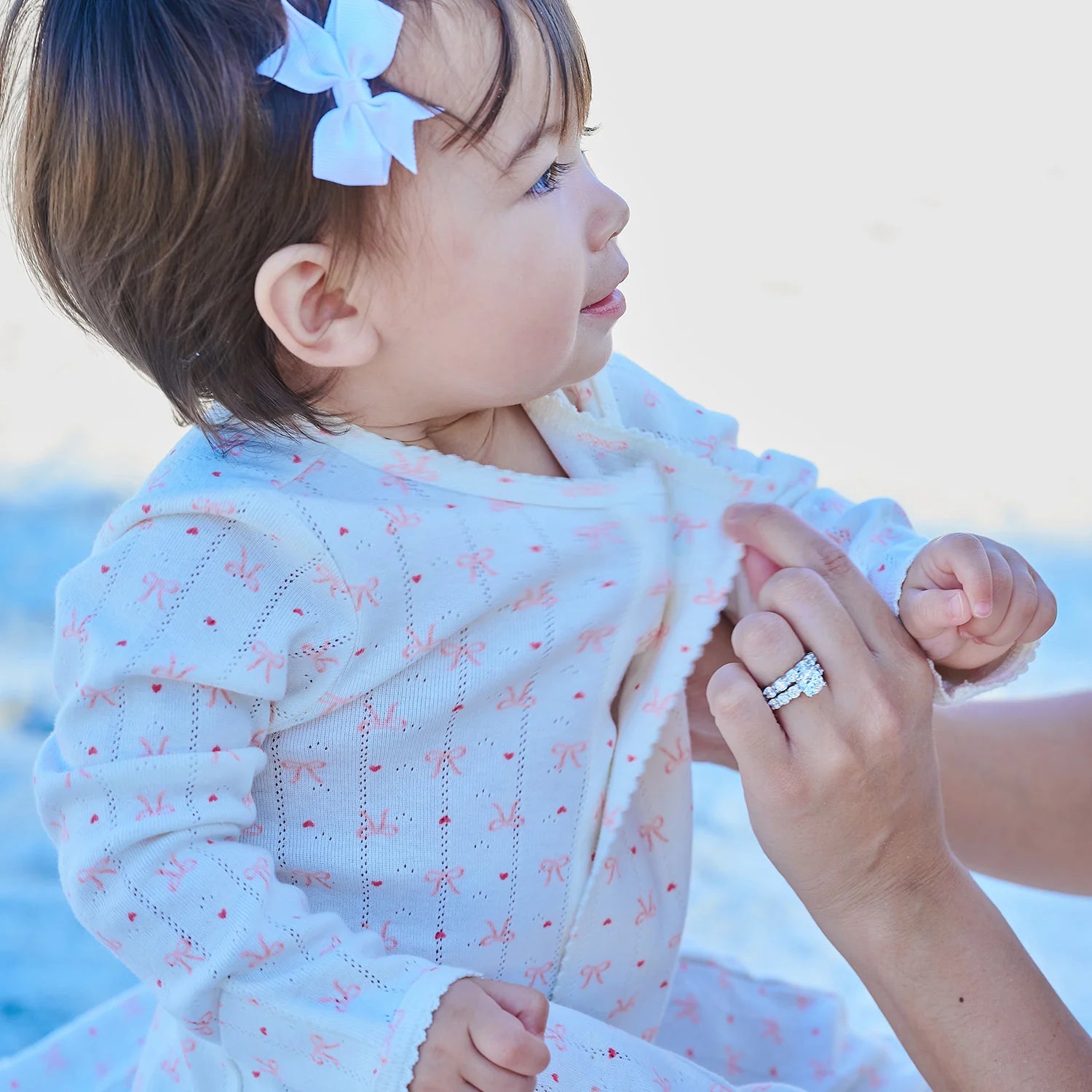 Child wearing a white outfit with pink patterns, held by an adult against a blurred natural background