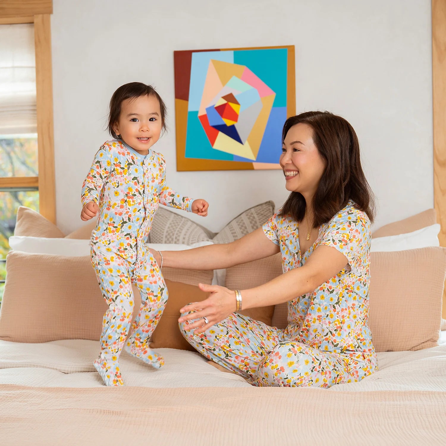 Woman and child in matching floral pajamas on a bed with a colorful abstract painting in the background.