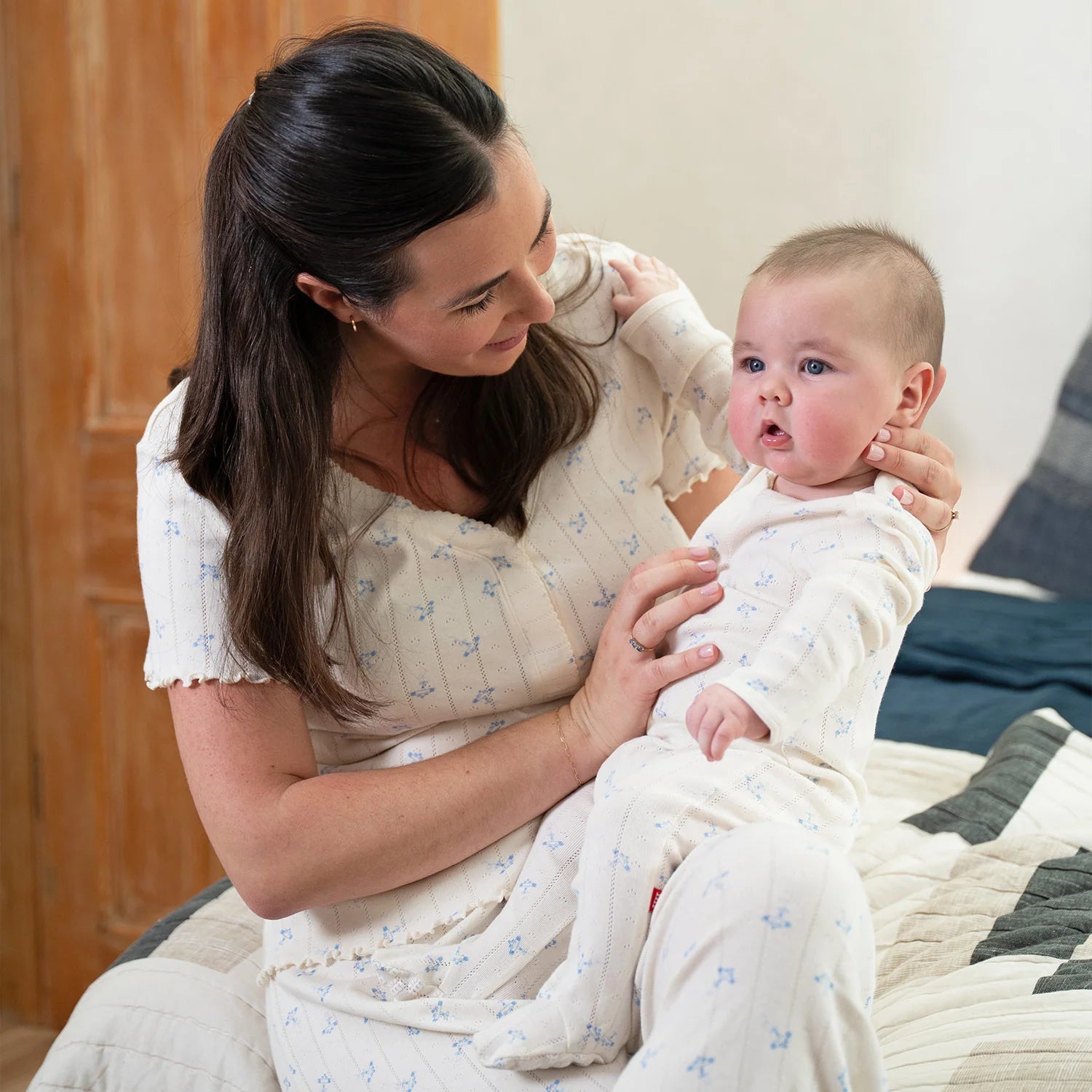 Woman holding a baby wrapped in a white blanket with blue patterns, sitting on a couch.