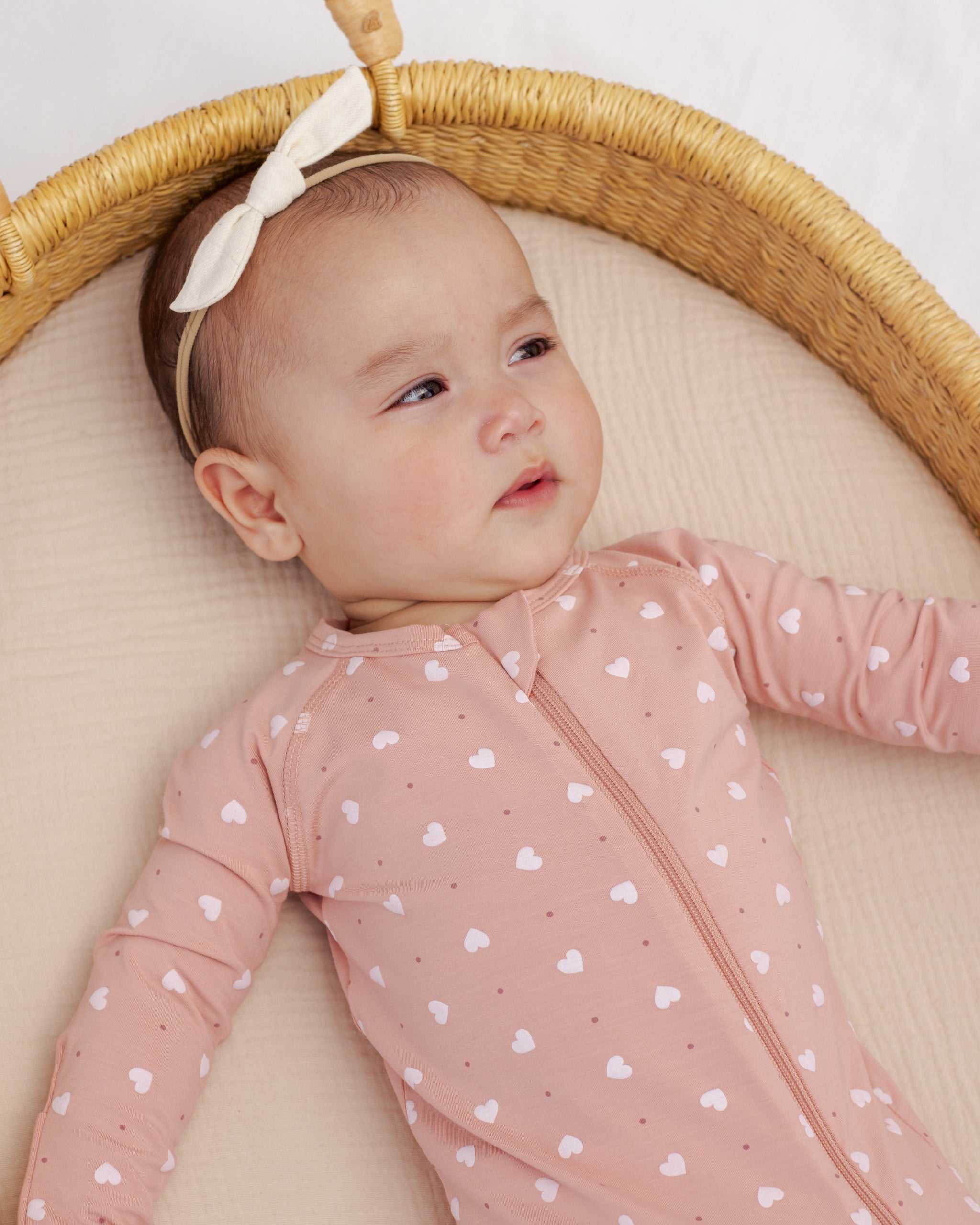 Baby in a pink heart-patterned onesie lying in a wicker basket with a white background